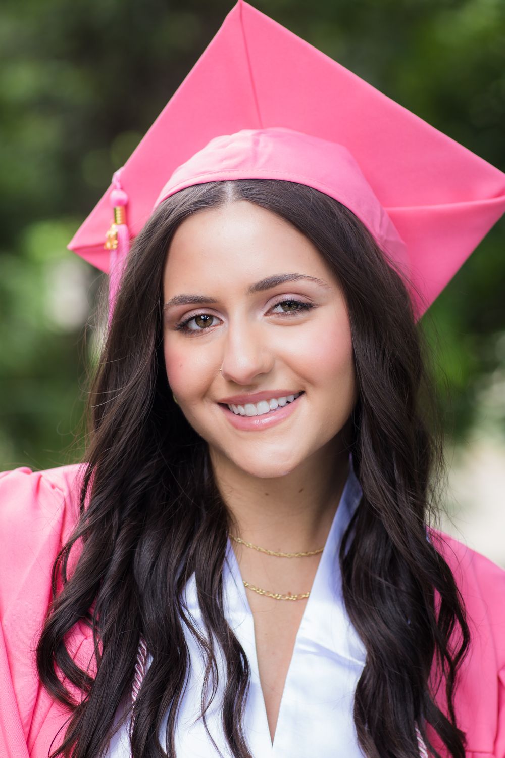 A graduation portrait featuring bright pink cap and gown against green foliage.