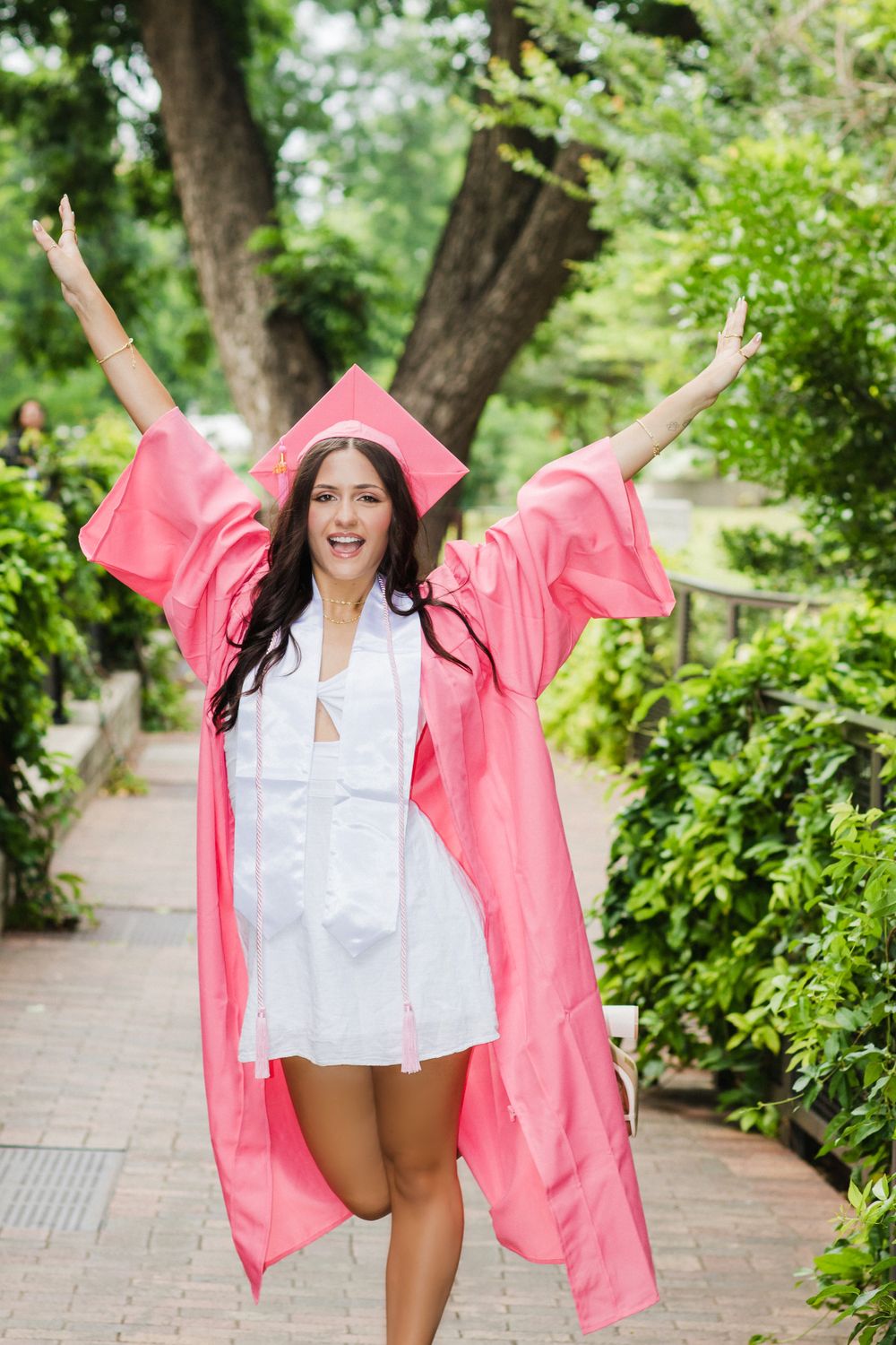 Graduate in pink cap and gown celebrates joyfully on a tree-lined pathway.