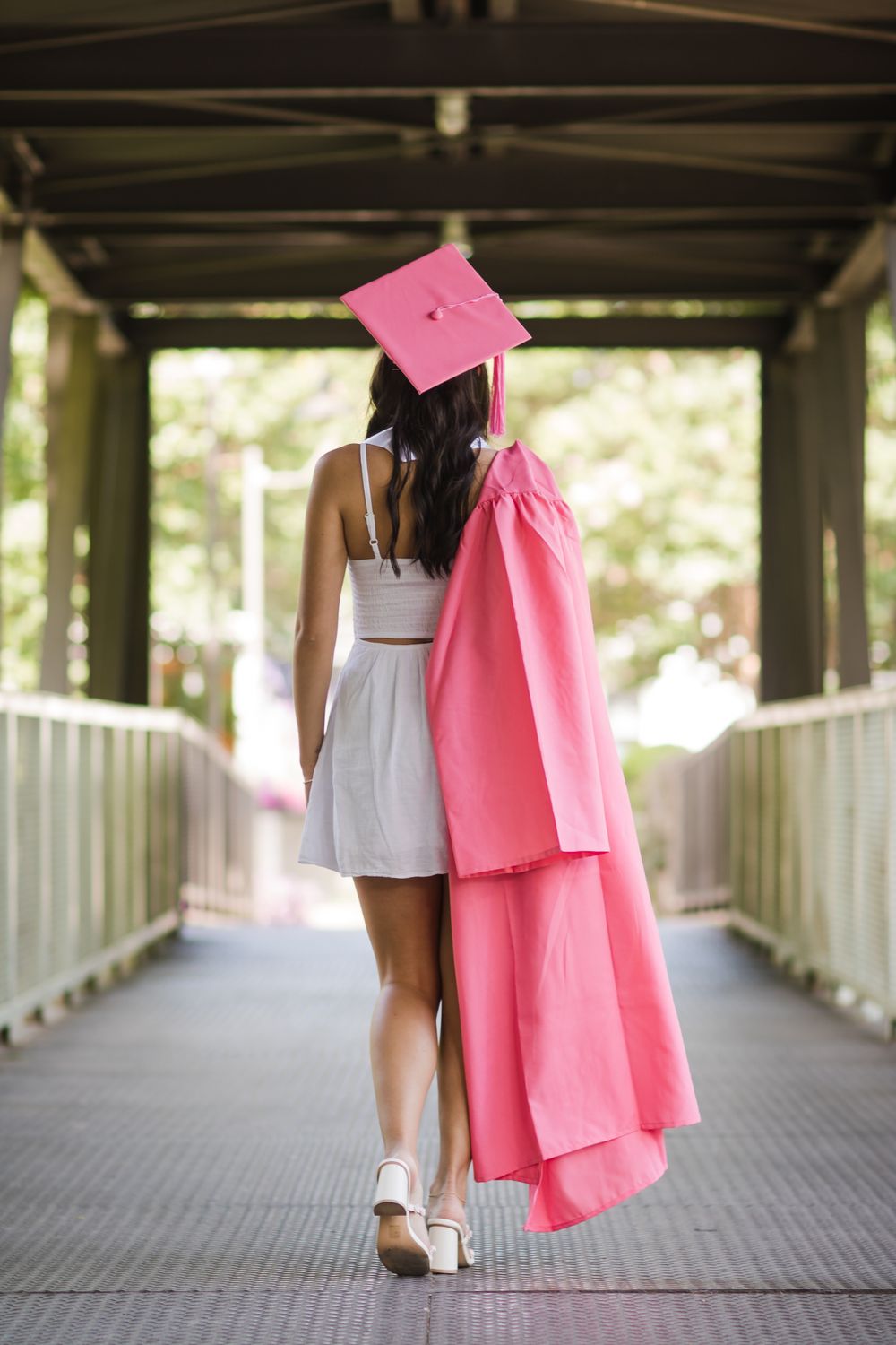 Graduate walking away on covered bridge wearing pink graduation gown and white dress.