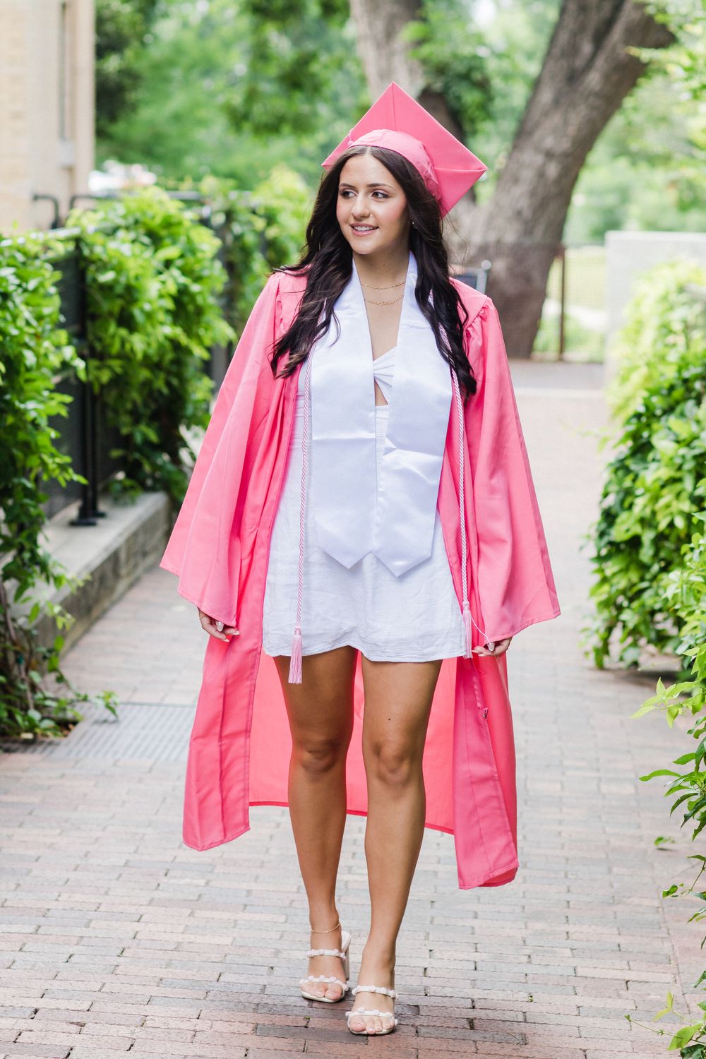 A graduate walks confidently down a brick path in pink graduation regalia.