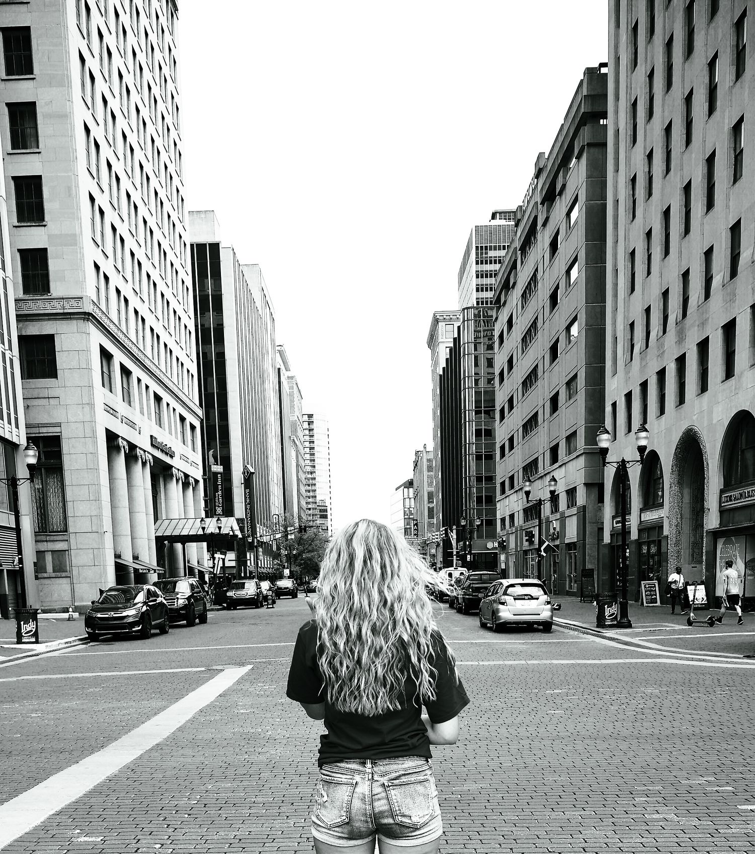 Back view of woman with long blonde hair looking down city street between tall buildings.