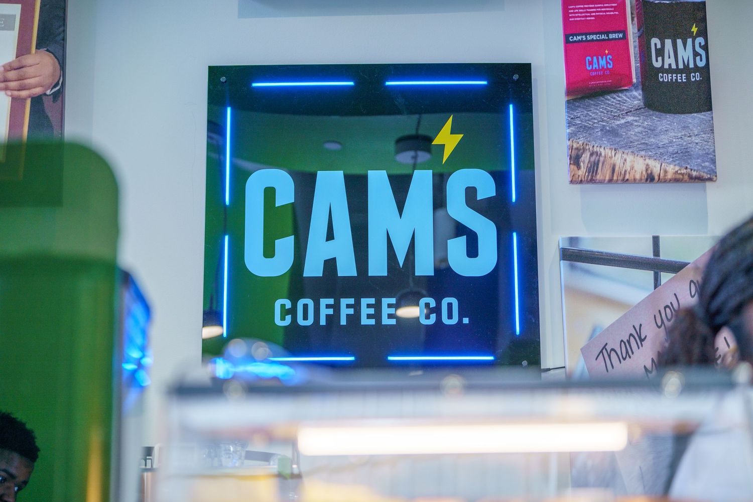 Illuminated blue neon sign for CAMS Coffee Co with lightning bolt logo on dark wall.