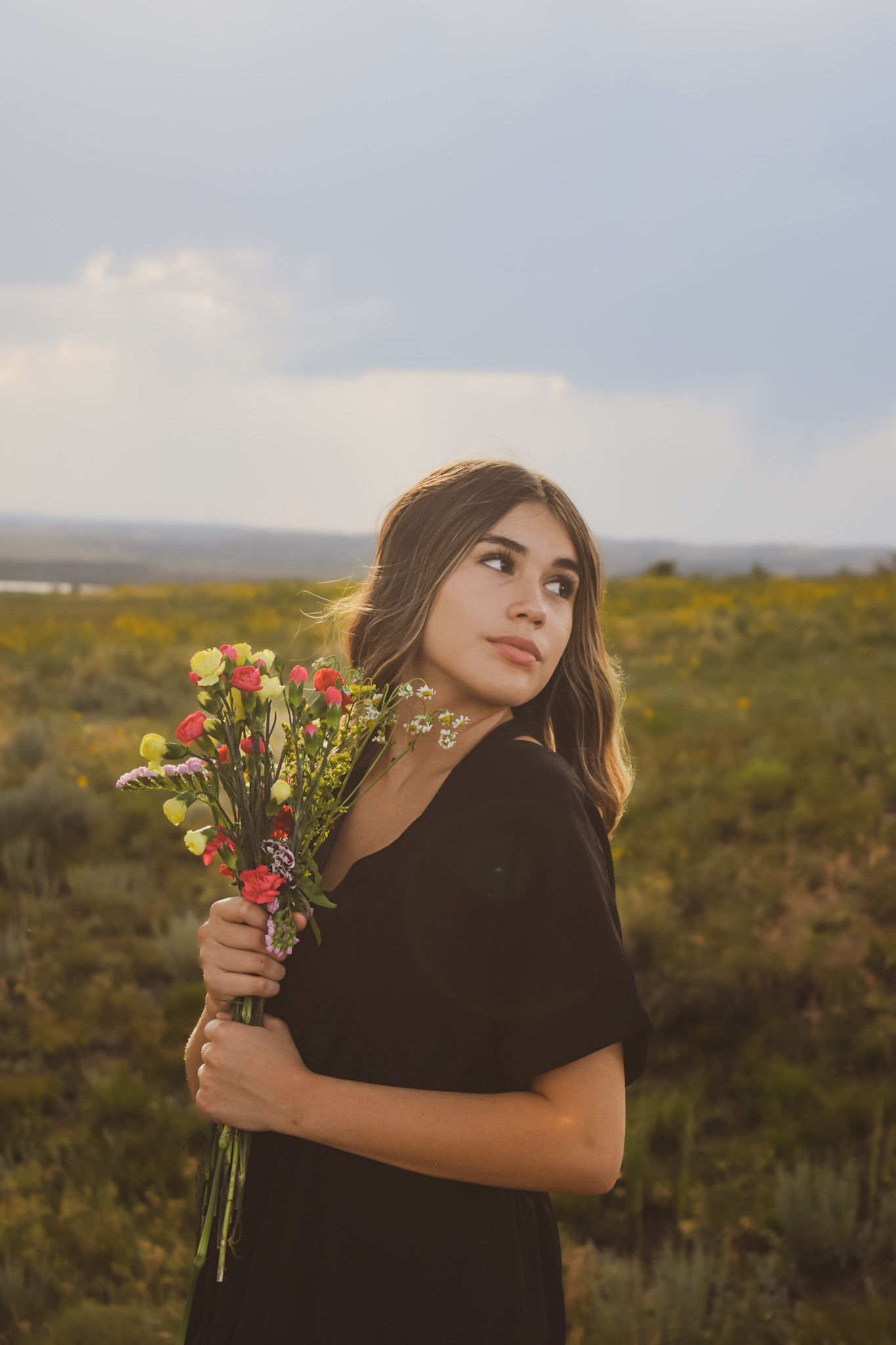 Person in black dress holding fresh flower bouquet against sunset landscape.