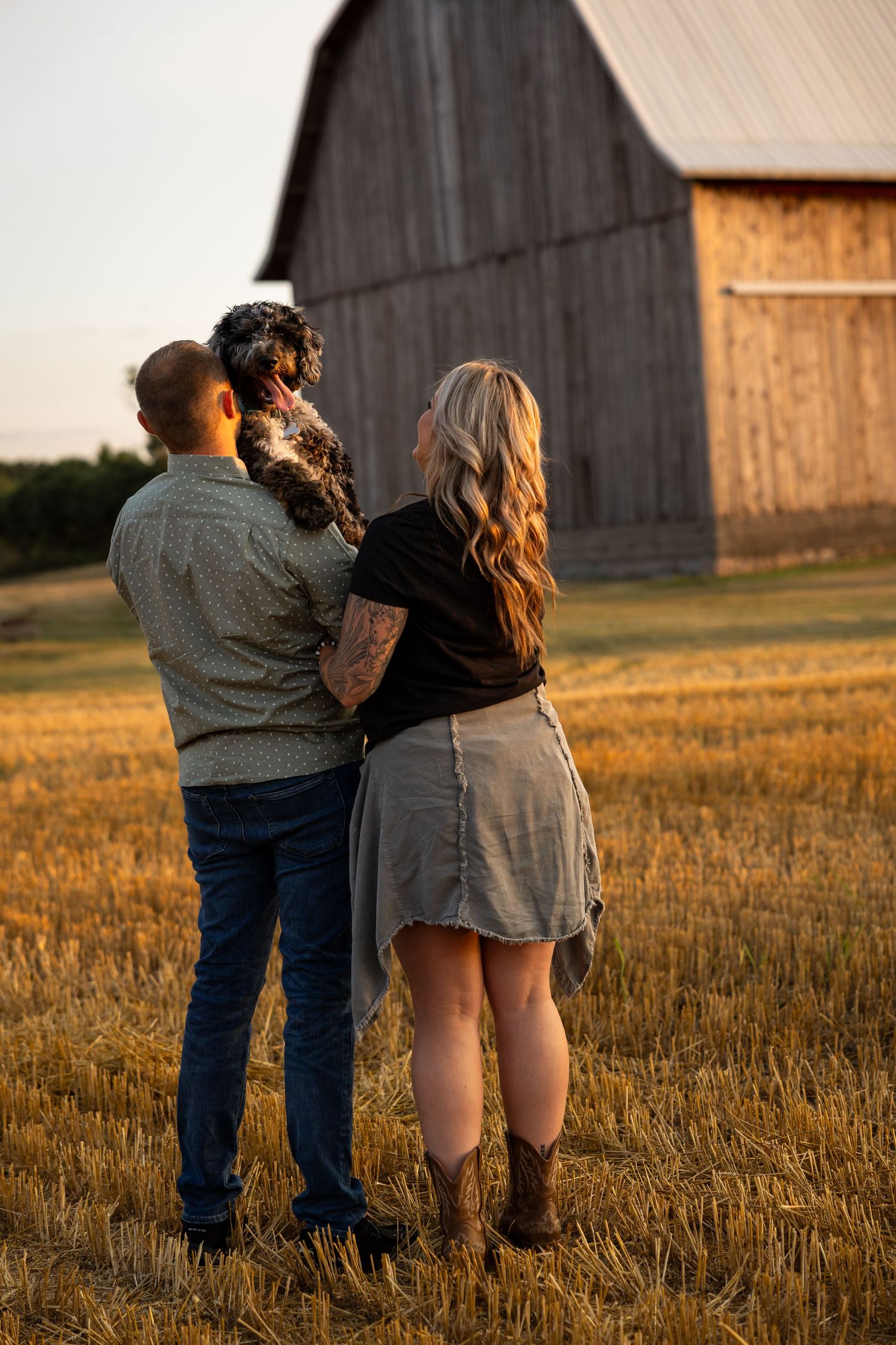 Couple holding a dog standing in a golden field facing a rustic wooden barn at sunset.