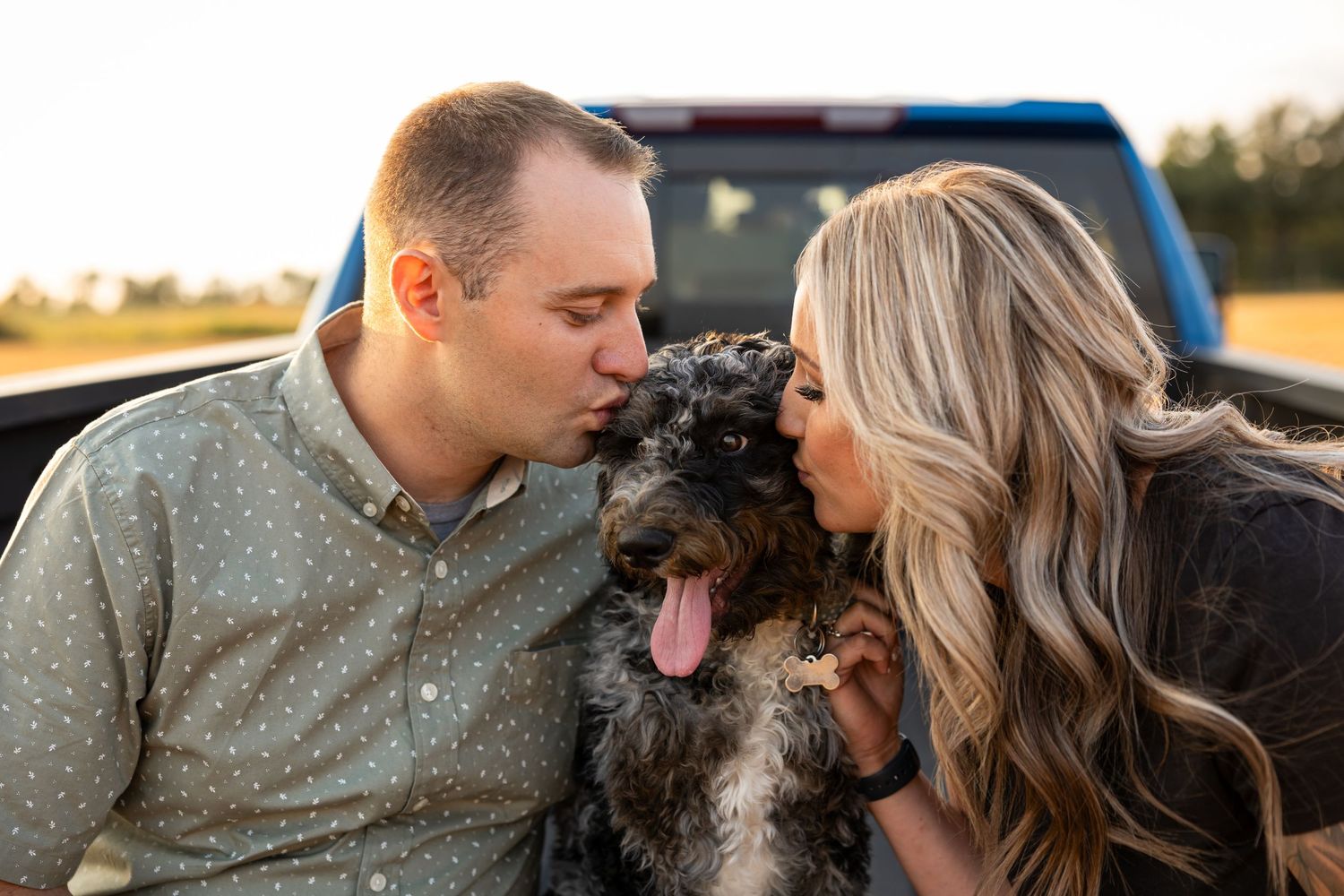 Couple kissing their doodle dog in a truck bed at golden hour.