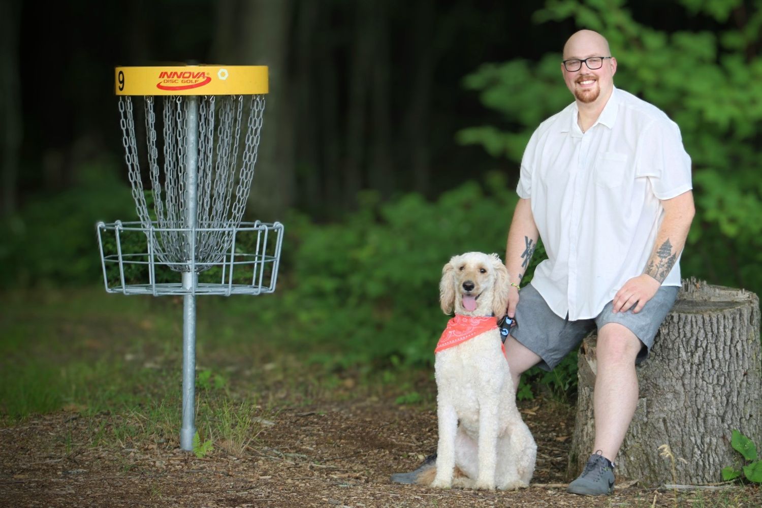 Man with tattoos sits on a log beside a golden doodle dog near a disc golf basket in a wooded park.