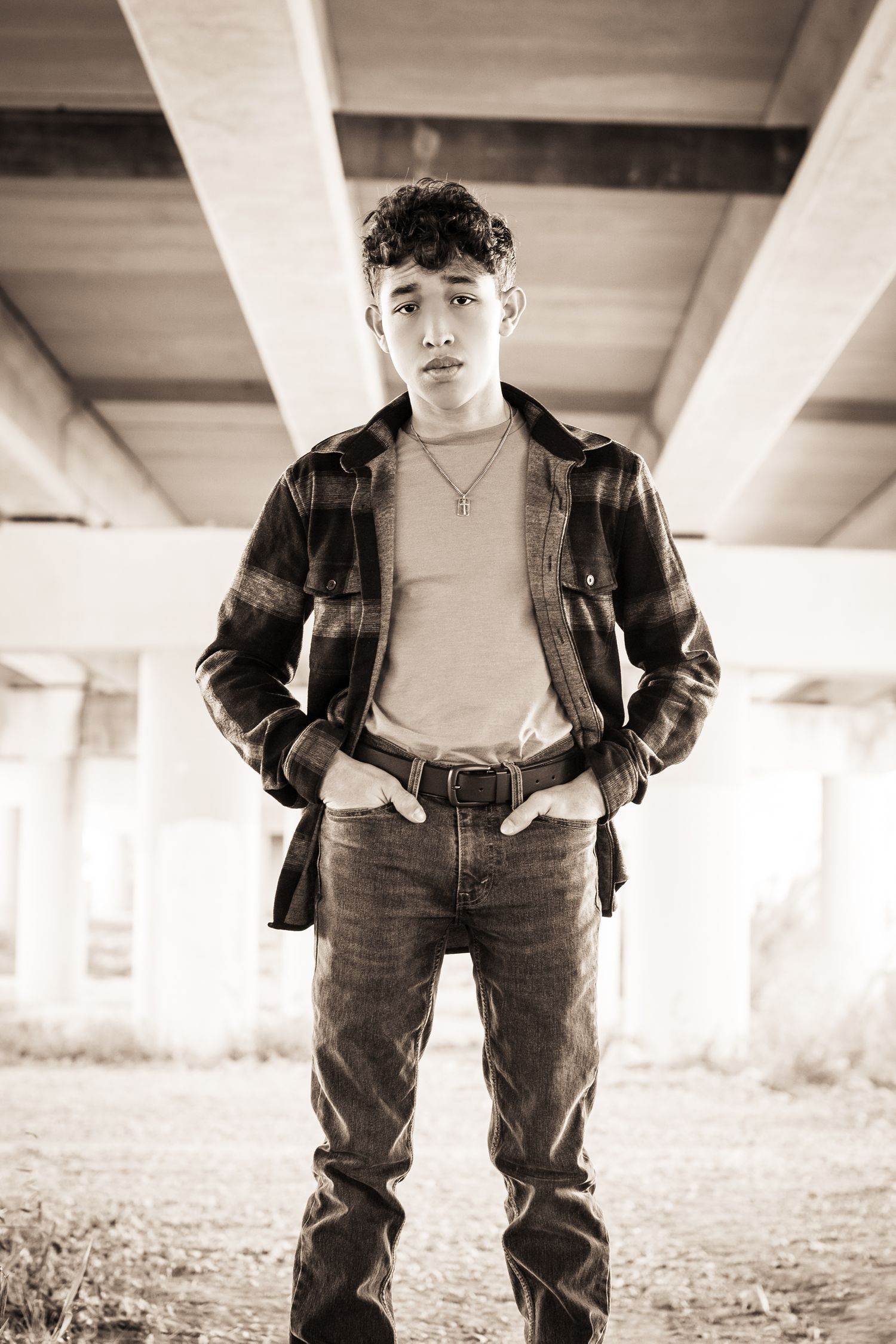 Black and white portrait of young person in plaid jacket and jeans under concrete structure.