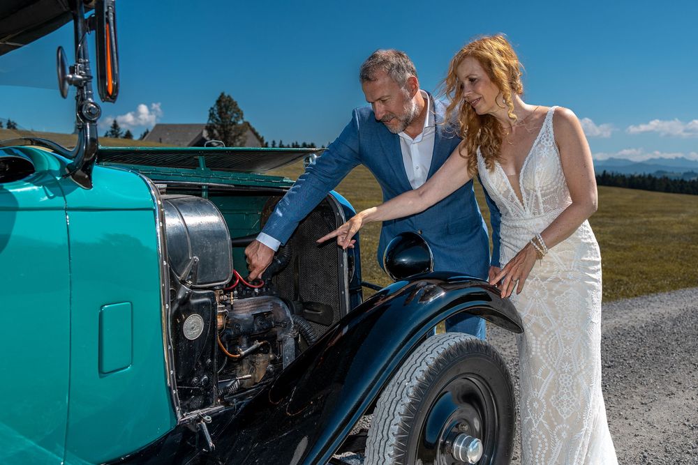 Bride and groom in elegant attire examining a vintage teal classic car engine on a sunny mountain road.