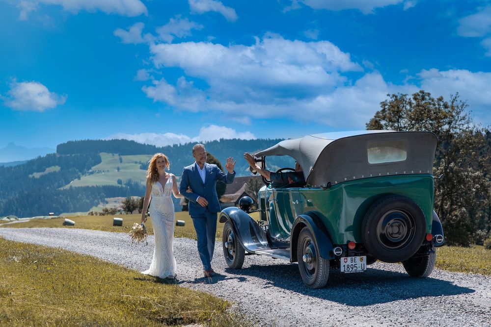 Bride and groom walk beside a vintage green car on a scenic mountain road under a bright blue sky.