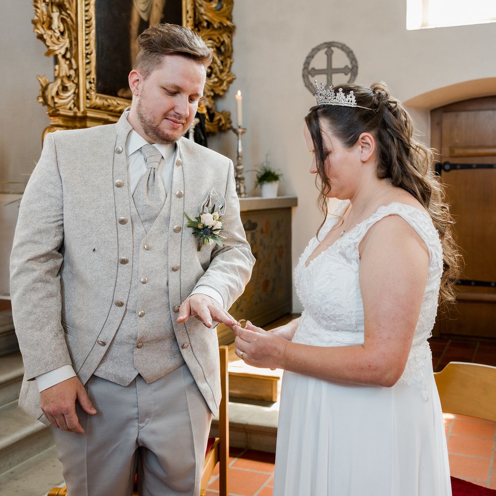 Bride placing wedding ring on groom's finger during church ceremony, both dressed in elegant wedding attire.