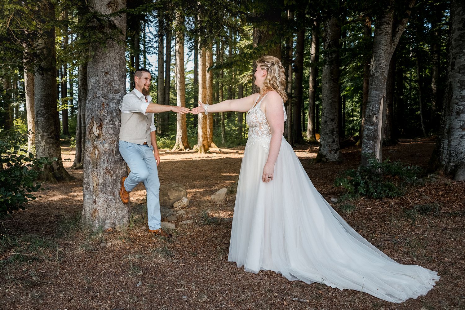 Bride in white gown and groom in light suit hold hands dancing in a sunlit forest wedding photo.