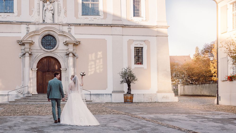 A wedding couple poses in front of a white church facade with elegant architectural details and arched windows.