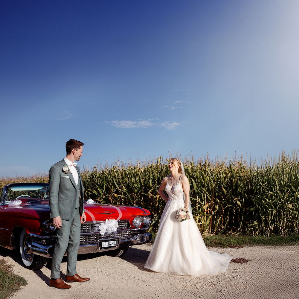Wedding party poses with vintage red car near cornfield on sunny day.