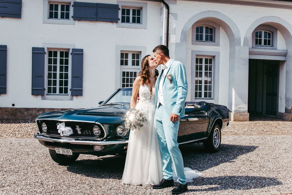 A vintage Ford Mustang parked in front of an elegant white mansion with shuttered windows.