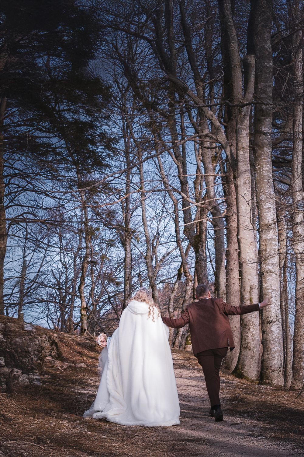 Couple walking through tall trees on forest path during wedding photoshoot.