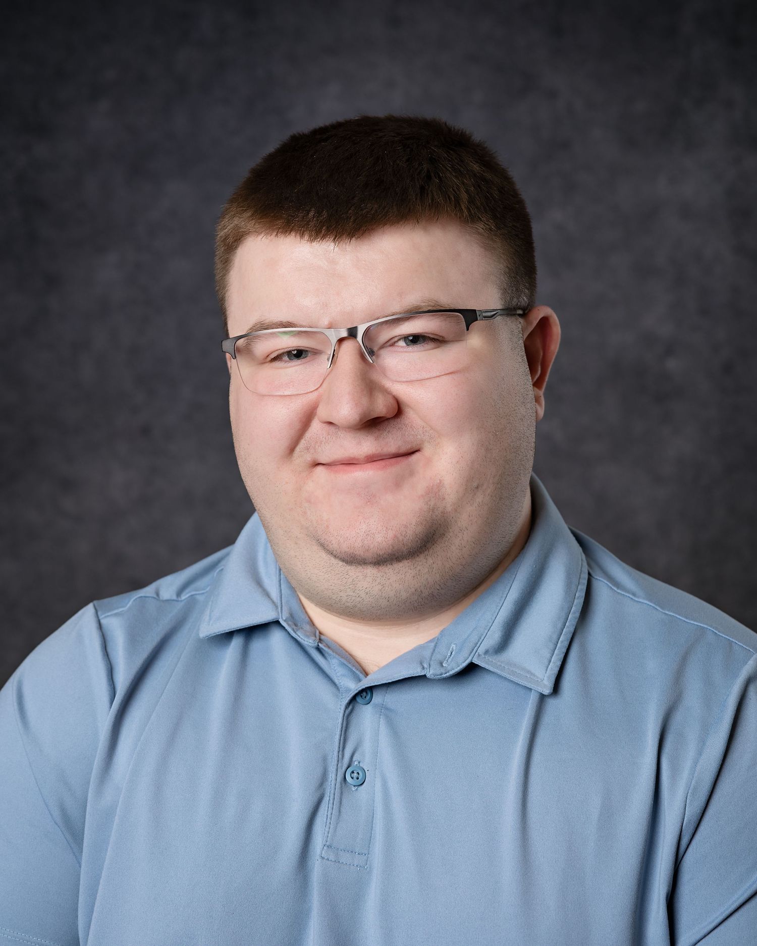 Young man with glasses wearing a light blue polo shirt poses for a professional headshot photo.
