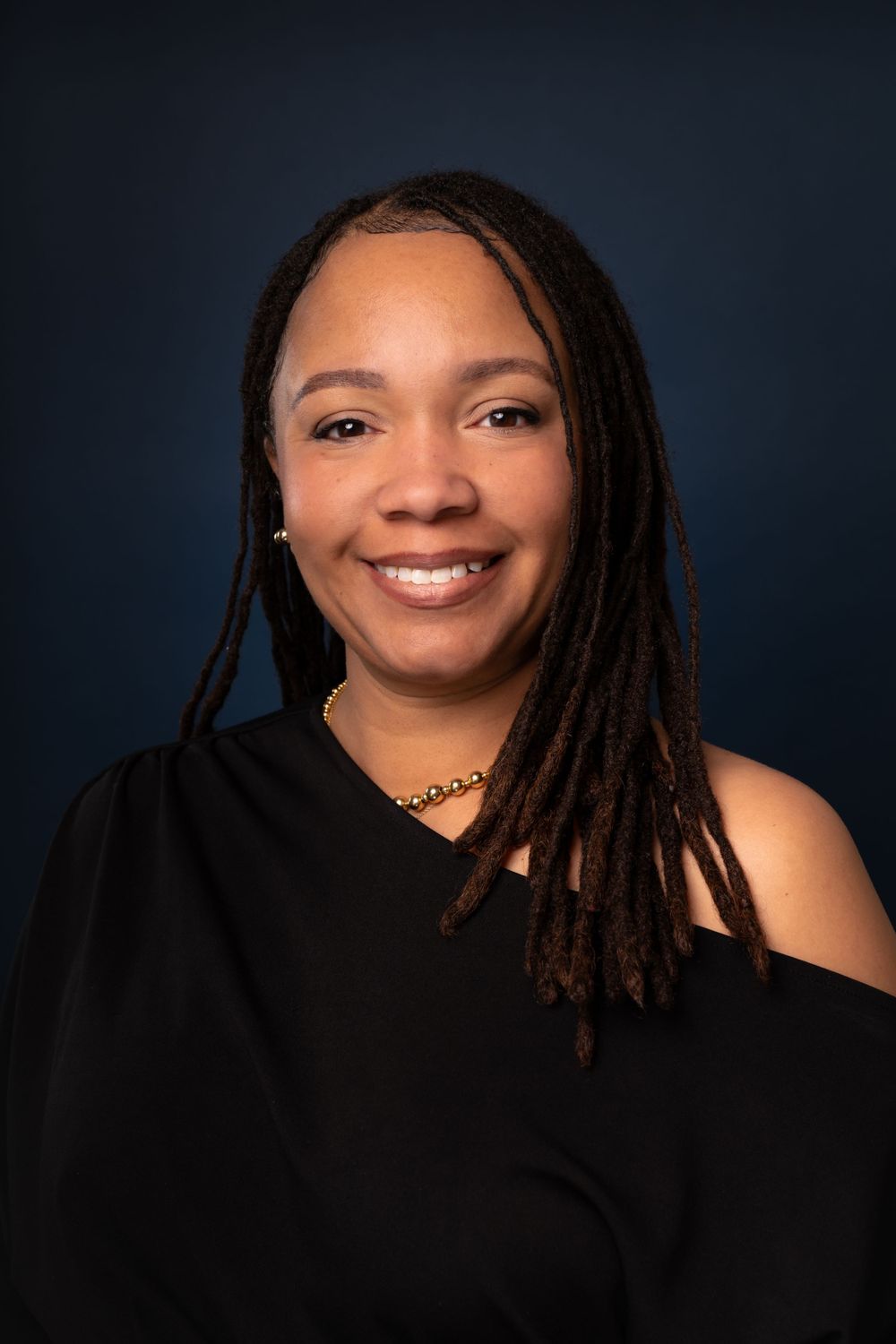 Portrait of person with dreadlocks wearing black top against dark studio backdrop.
