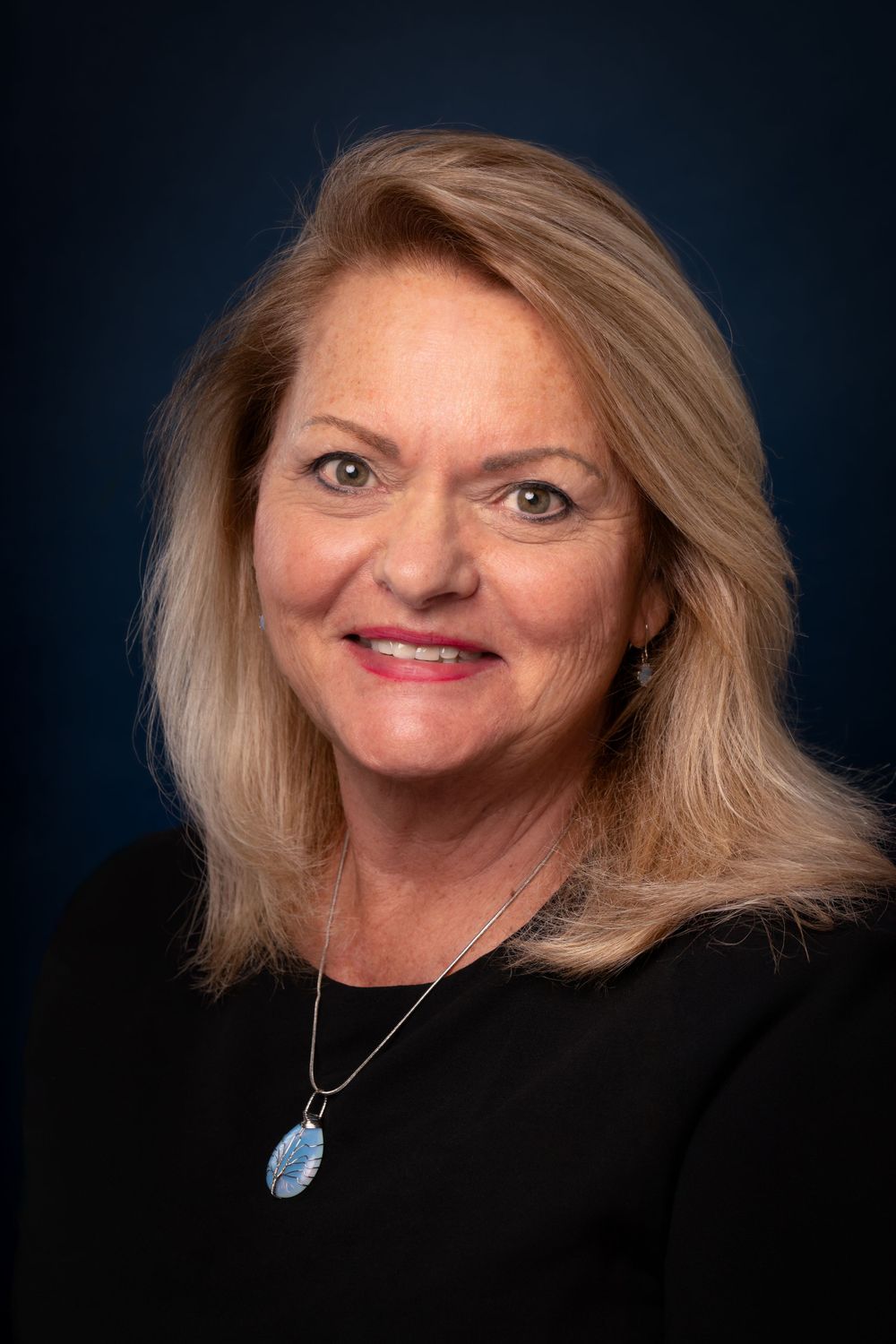Professional headshot of person with blonde hair wearing black top against dark background.