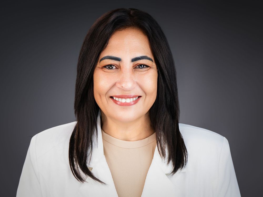 Professional headshot of a smiling woman in a white blazer against a dark gray background.