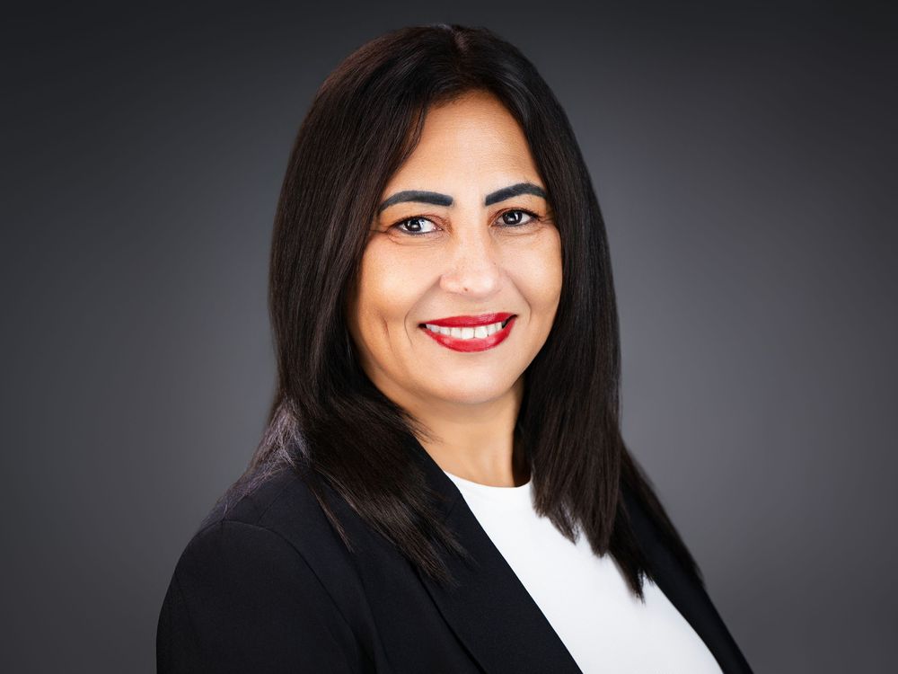 Professional headshot of a smiling woman with dark hair wearing a black blazer and white top against a gray background.