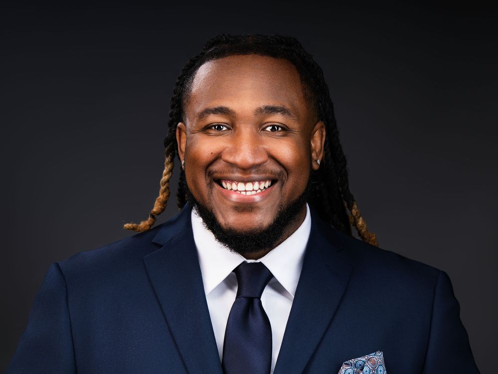 Professional headshot of a smiling man in a navy suit with tie and pocket square, wearing locs hairstyle.