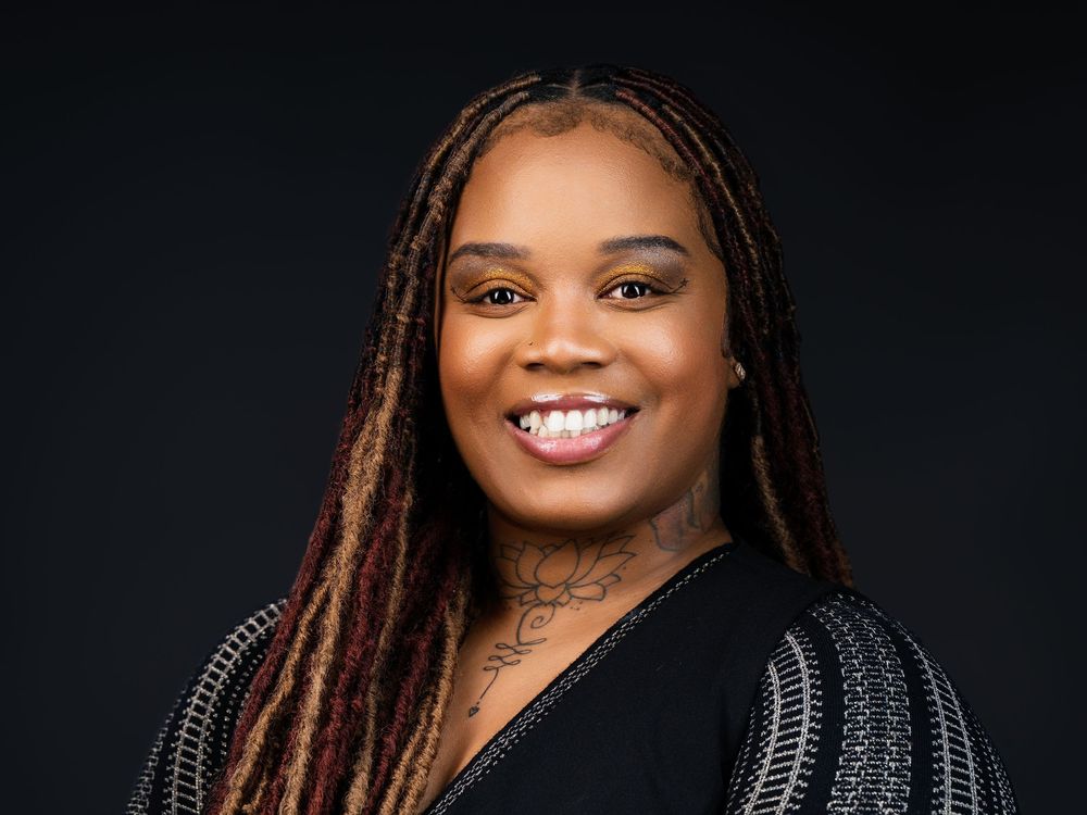 Smiling woman with long braids and neck tattoo wearing a black top against a dark background.