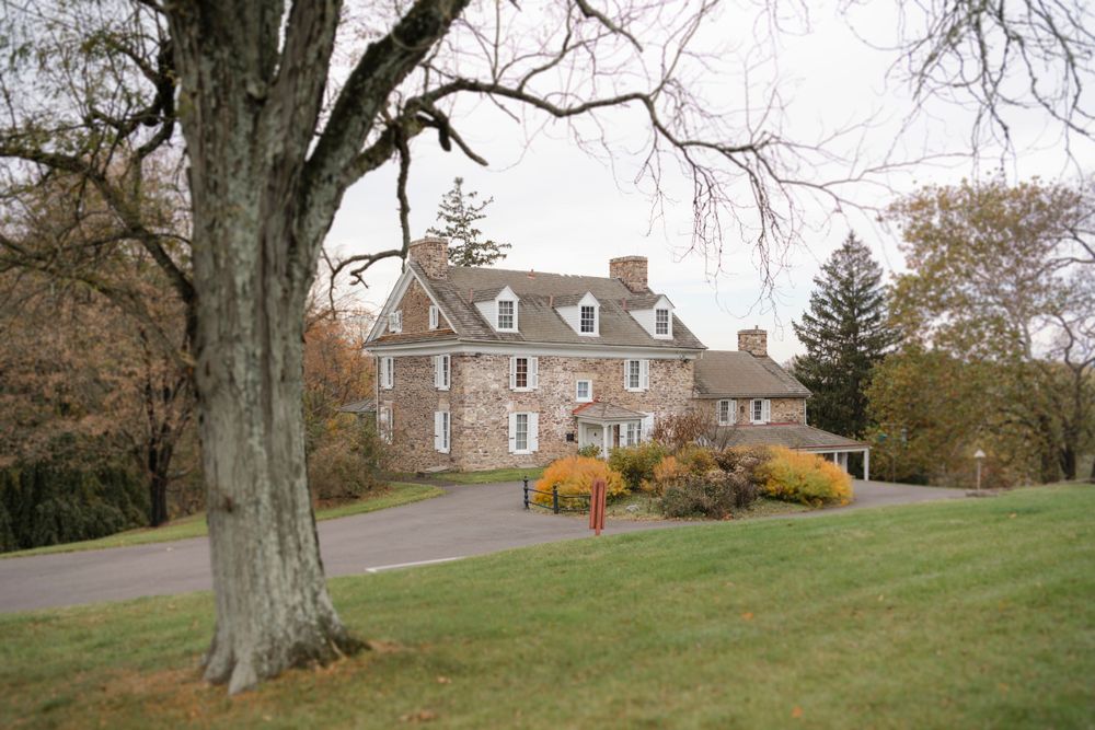 Large colonial stone house with dormer windows sits on a grassy hill surrounded by tall trees in autumn.