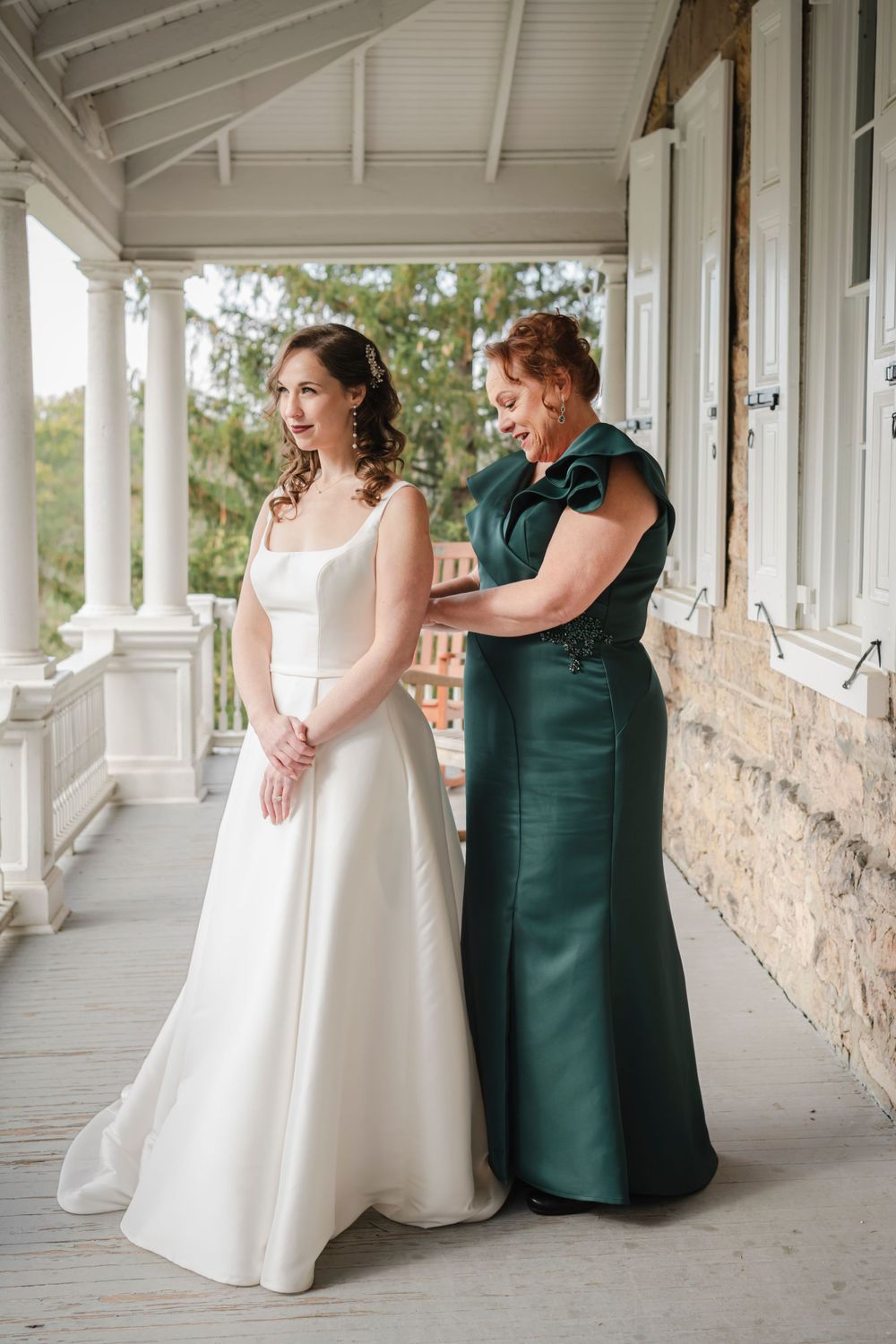A bride in a white wedding gown getting help with her dress from her mother wearing an emerald green formal dress on a porch.