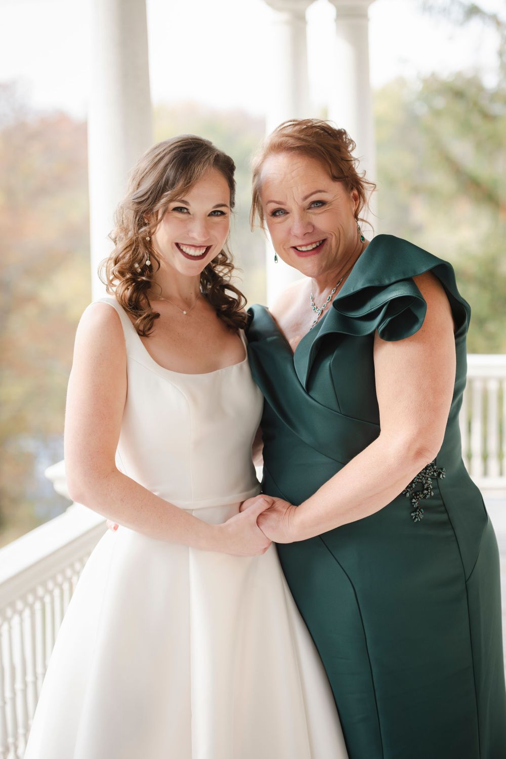 Bride and her mom in formal attire, one wearing a white wedding dress and one in an elegant dark green dress, pose on a porch.