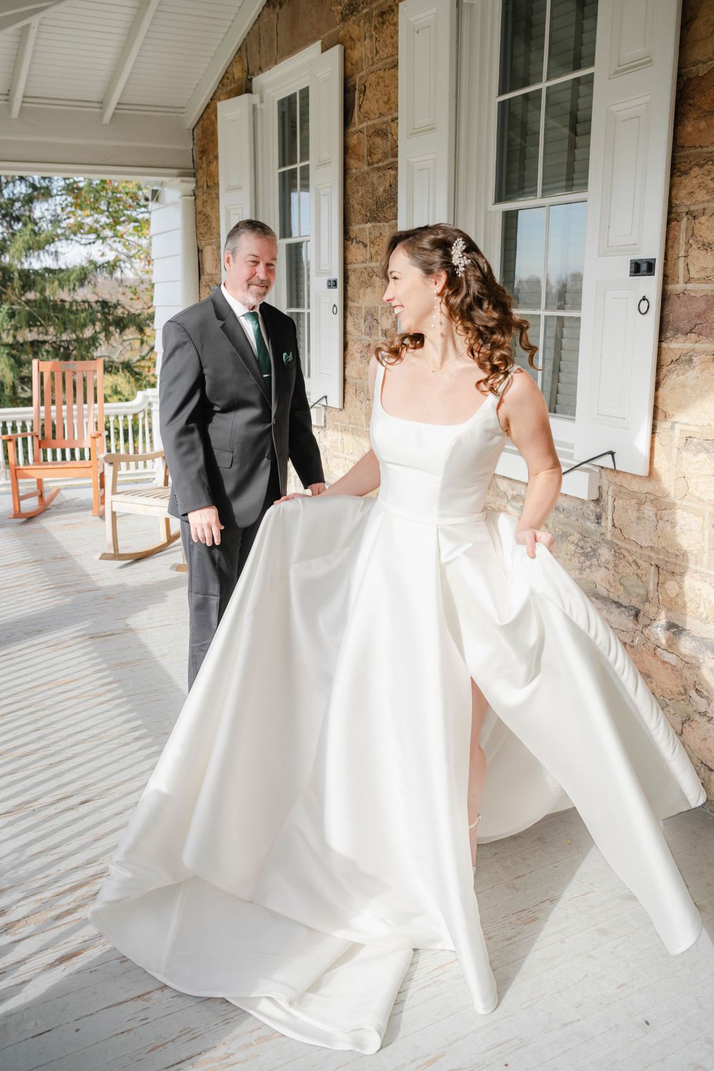 A bride in a flowing white gown twirls on a stone porch while dad in a dark suit looks on during a wedding day.
