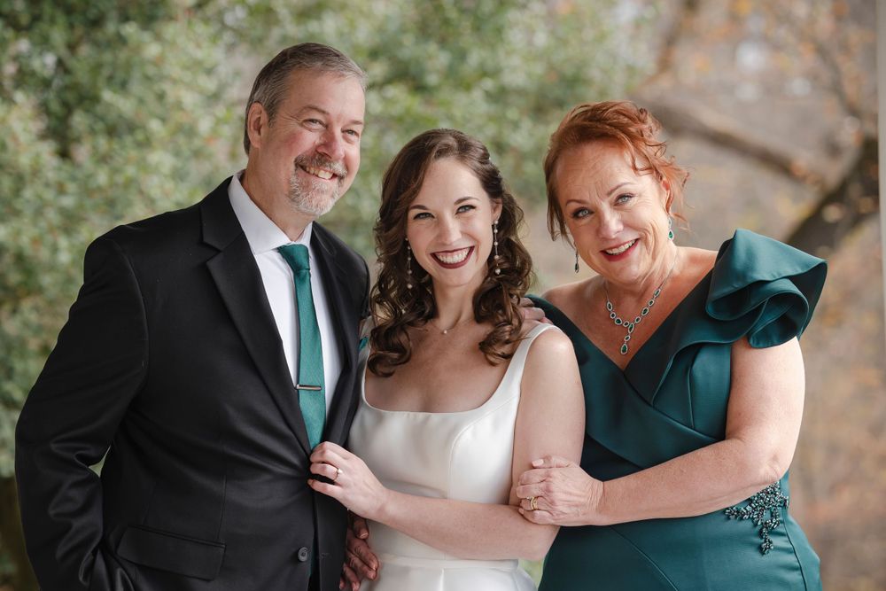 A bride and her parents pose together for a formal portrait outdoors, wearing a black suit, white dress, and emerald green formal gown.