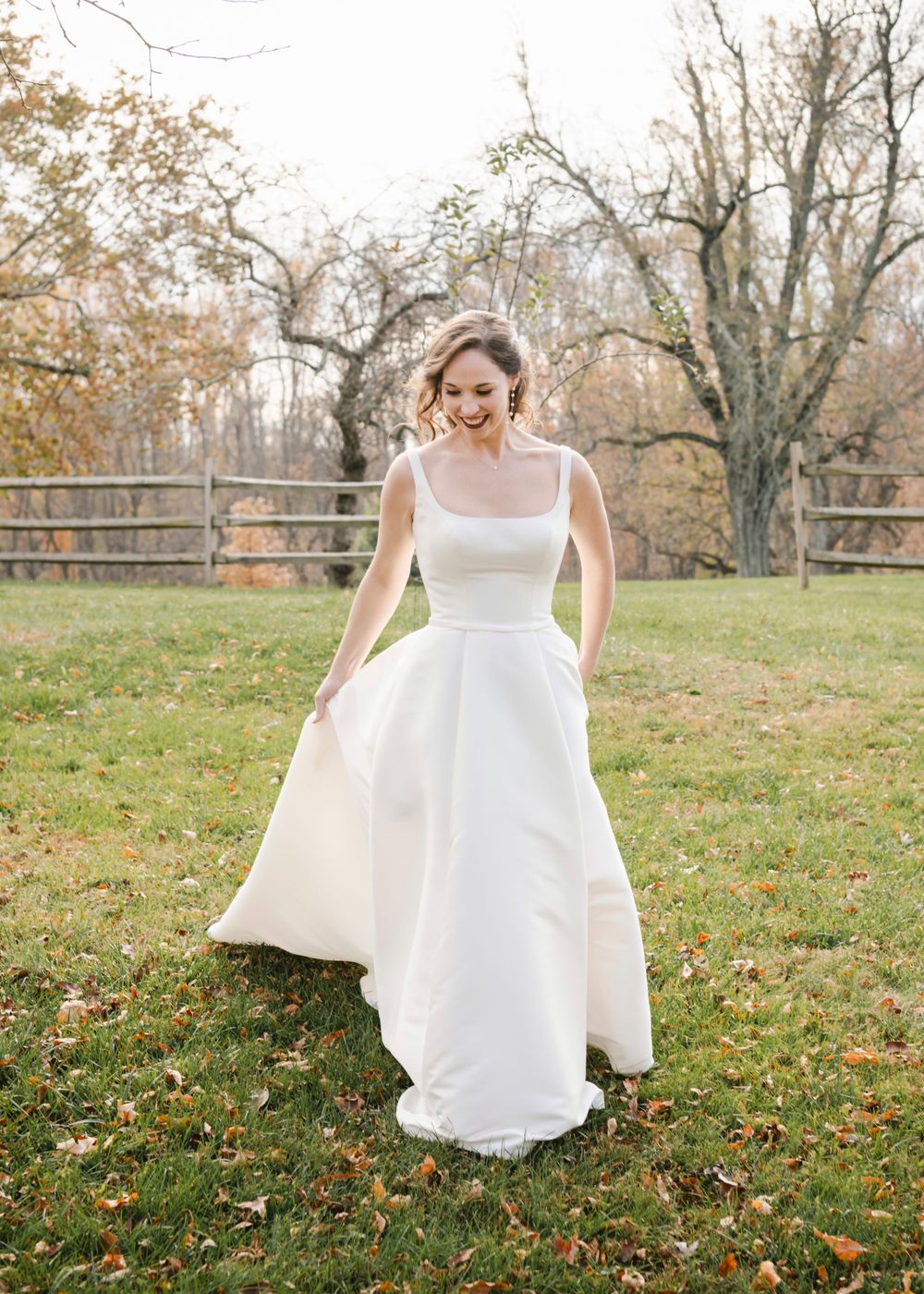 A bride in a classic white wedding gown with pockets walks in a rustic autumn lawn with bare trees and fallen leaves.