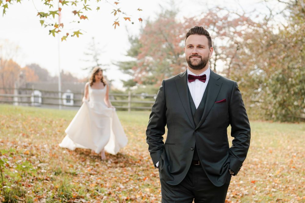 A happy couple in wedding attire prepare for the first look in a fall setting with autumn leaves scattered on the ground.