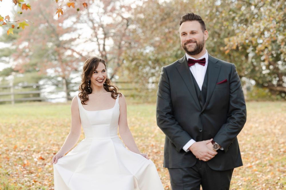 A couple in formal wedding attire prepares for the first look among autumn leaves in an outdoor setting.