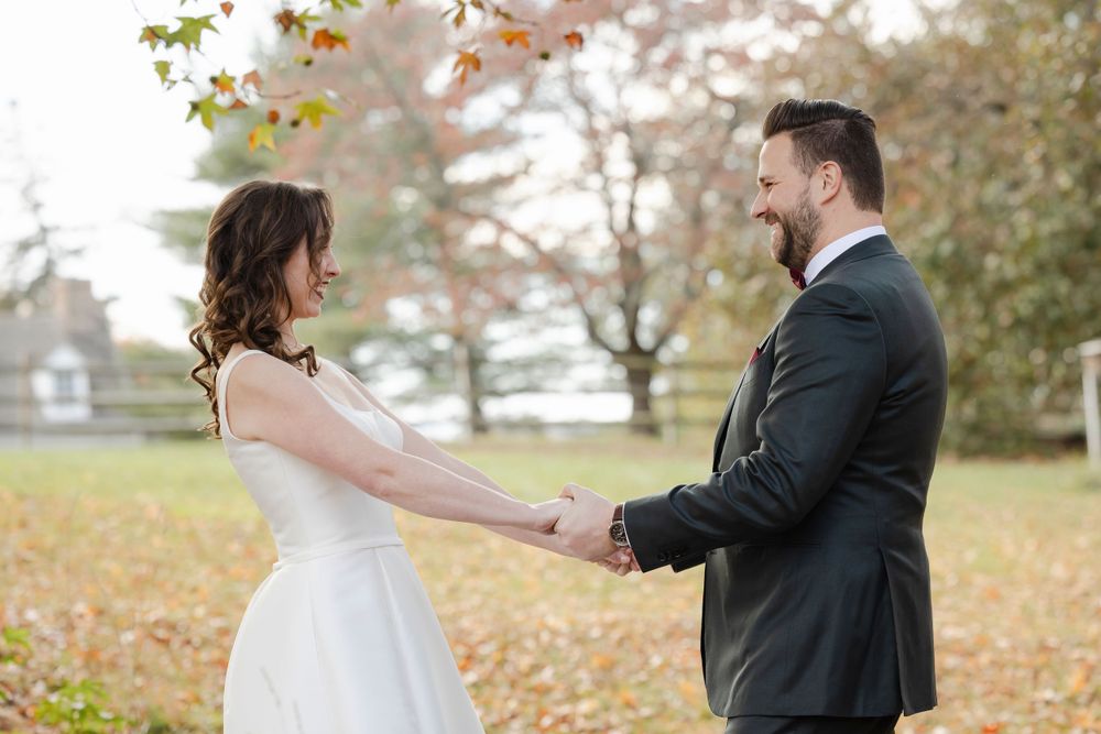 A bride and groom hold hands while standing face-to-face in a park during autumn with falling leaves around them.