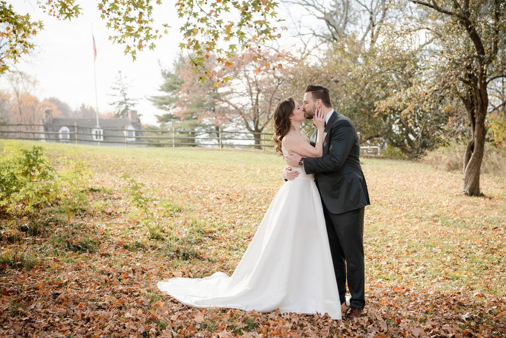 A romantic wedding couple shares an intimate moment surrounded by fall foliage in a park setting at sunset.