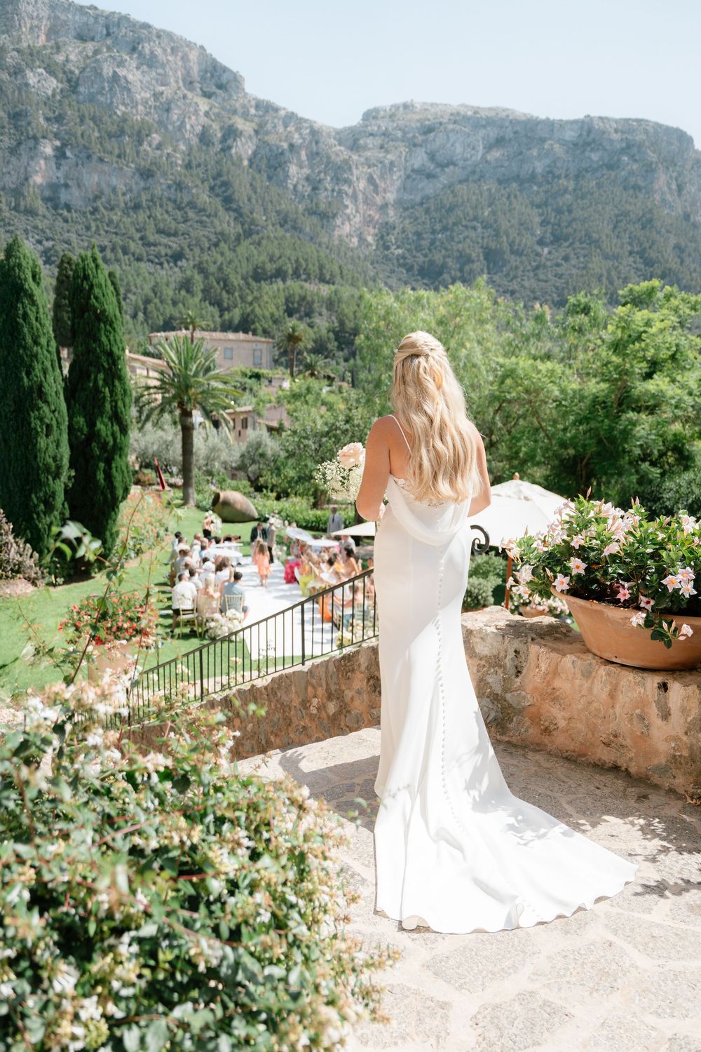 A figure in a white wedding dress stands on a scenic terrace overlooking mountains and gardens in Mallorca.