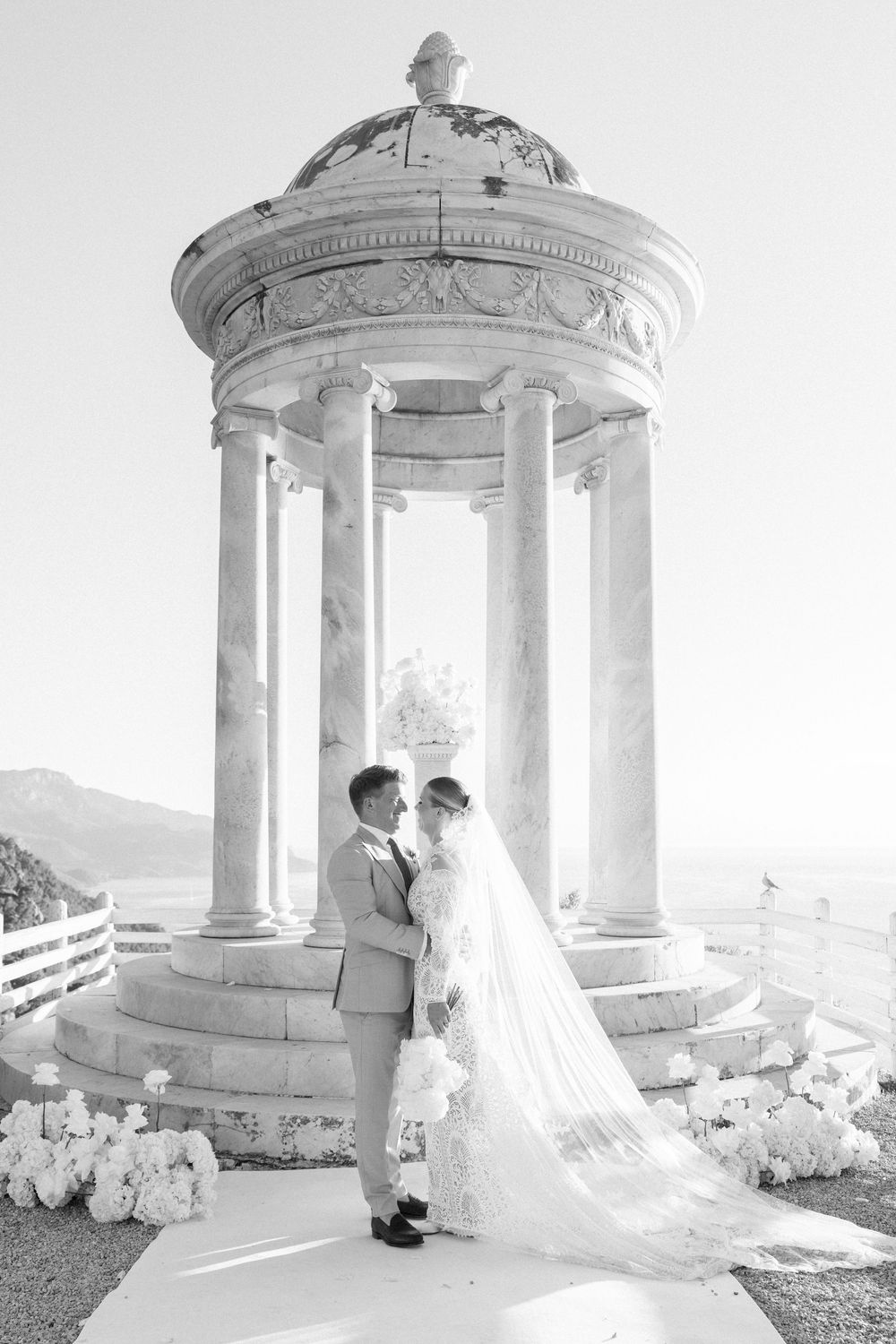 A romantic wedding photoshoot in front of an ornate white gazebo with flowing bridal veil and decorative white flowers.