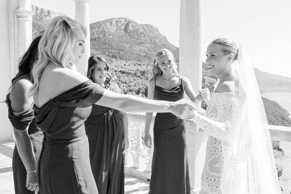 A black and white wedding photo showing a touching moment between bridesmaids and a bride on a balcony with mountains behind.