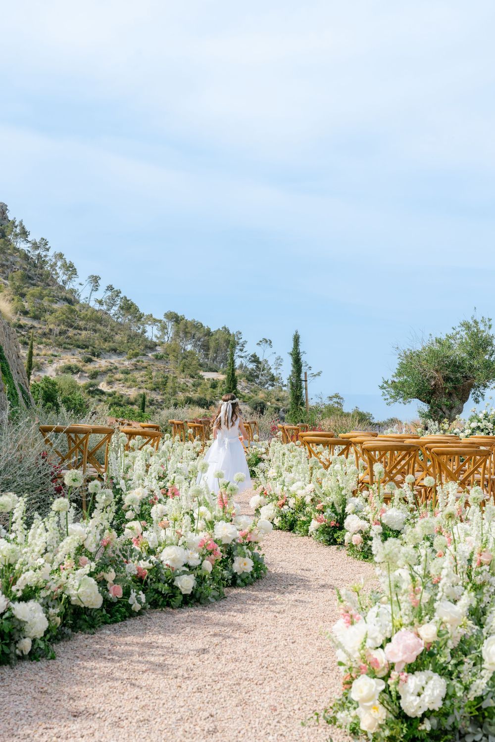 A garden path lined with lush white hydrangeas and roses leads to a wooden arch under a blue sky at an outdoor venue.