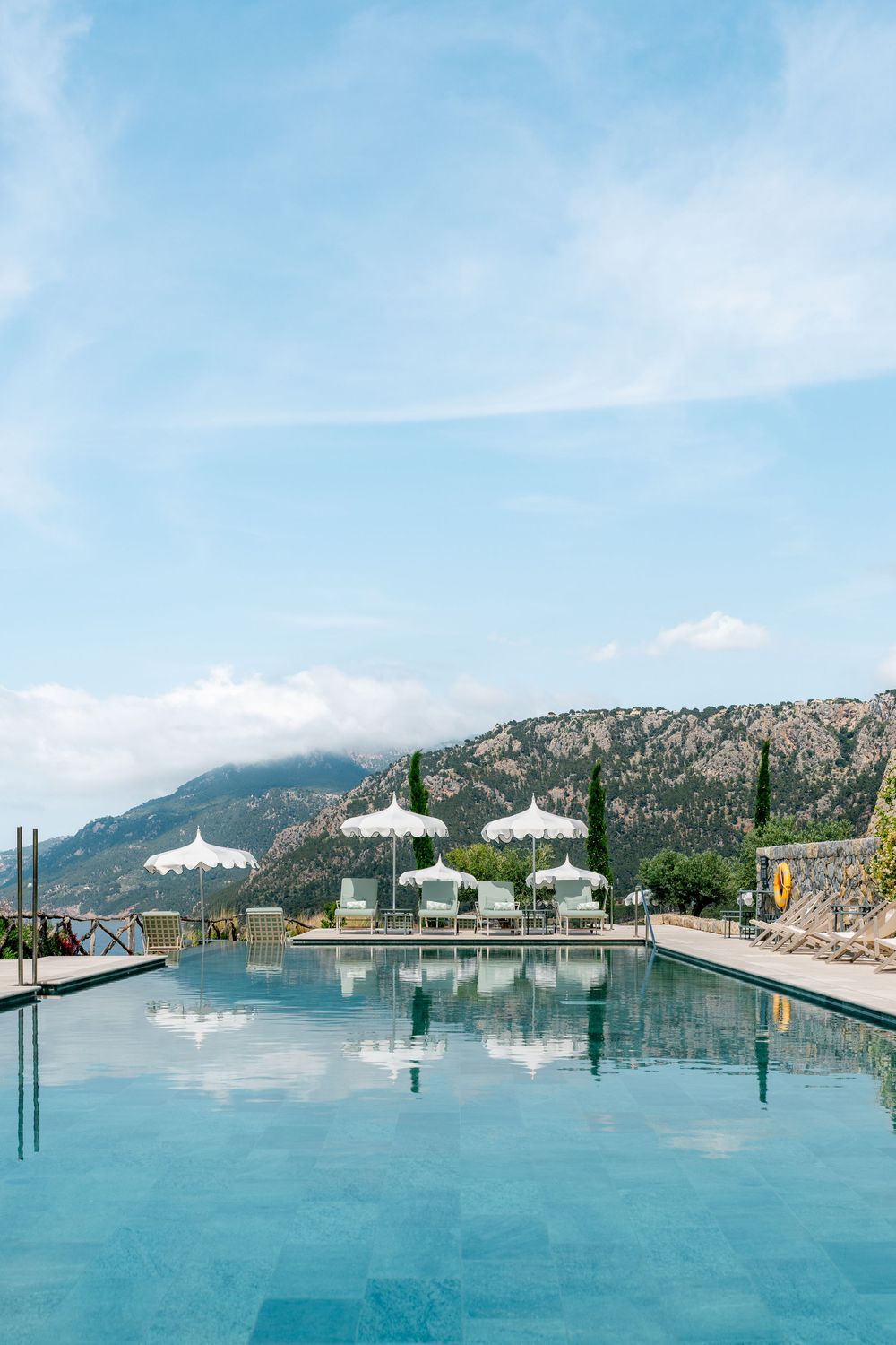 Panoramic view of a serene infinity pool overlooking mountains and ocean with white umbrellas and lounge chairs.