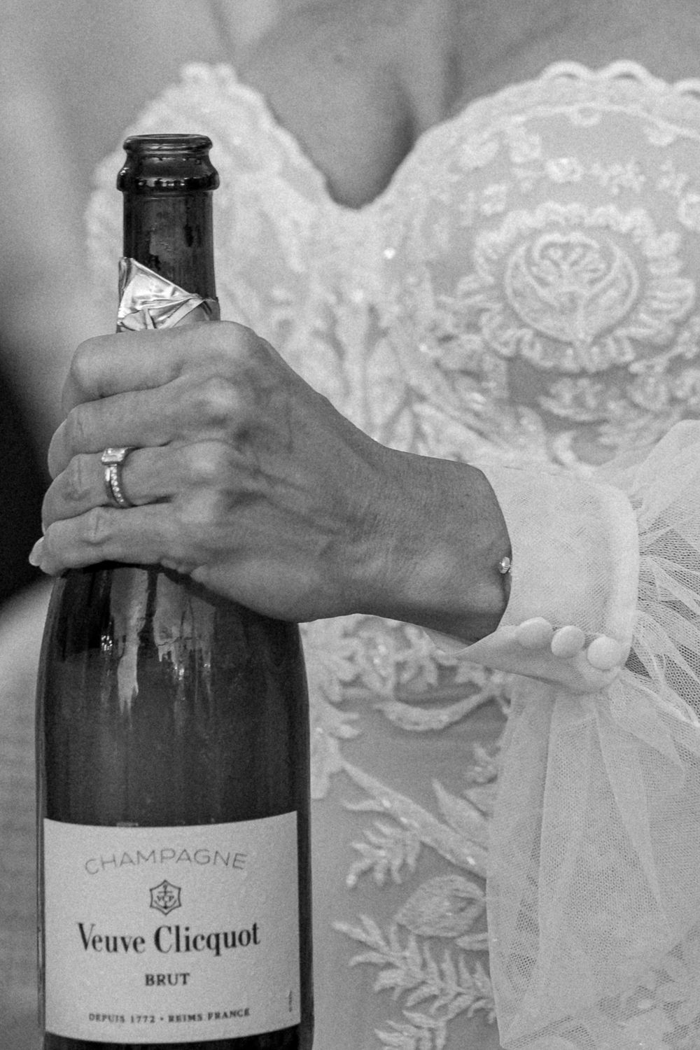 Close-up of wedding dress detail and hand holding Veuve Clicquot champagne bottle.