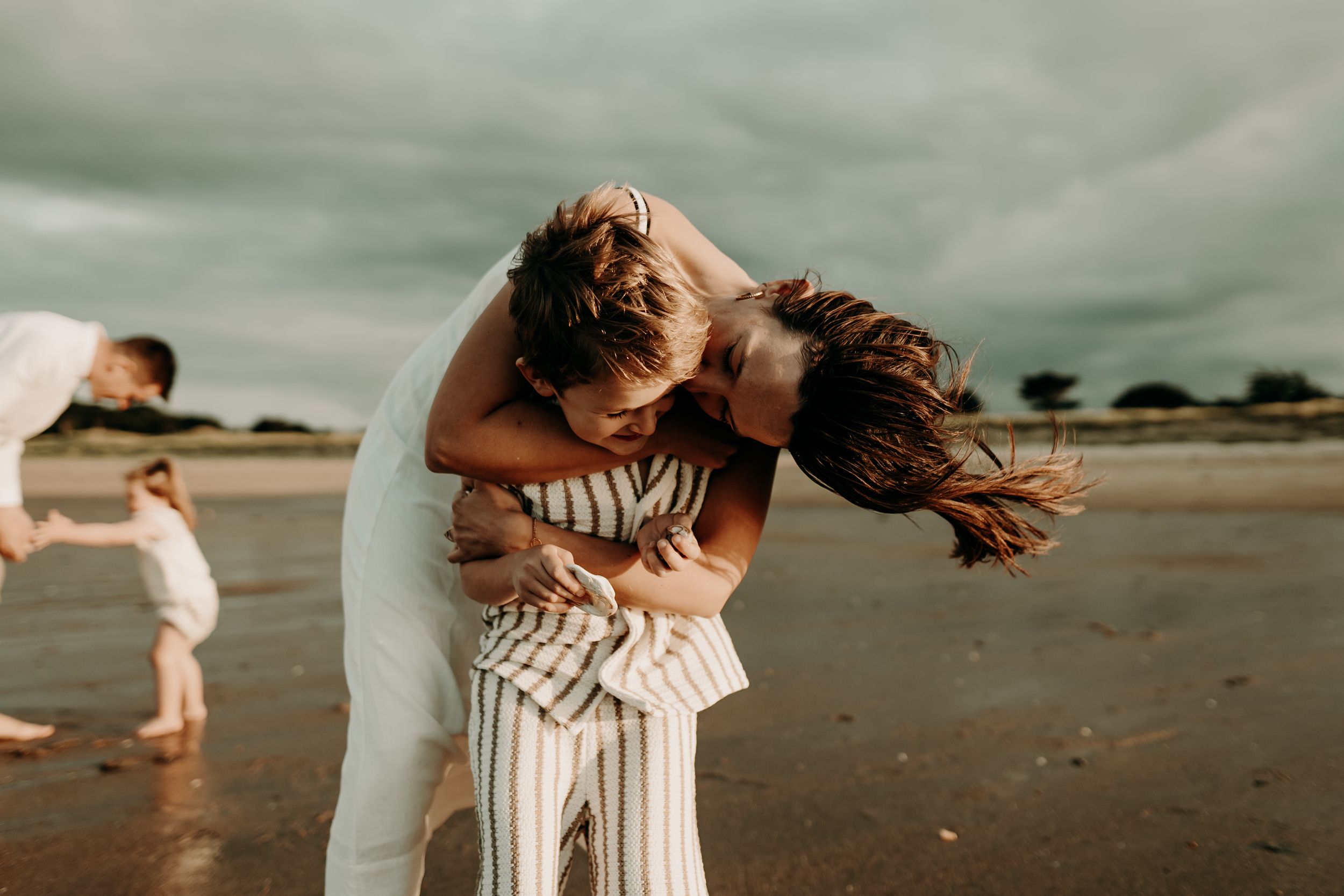 A couple embraces tenderly on a sandy beach at sunset while waves crash in the background.