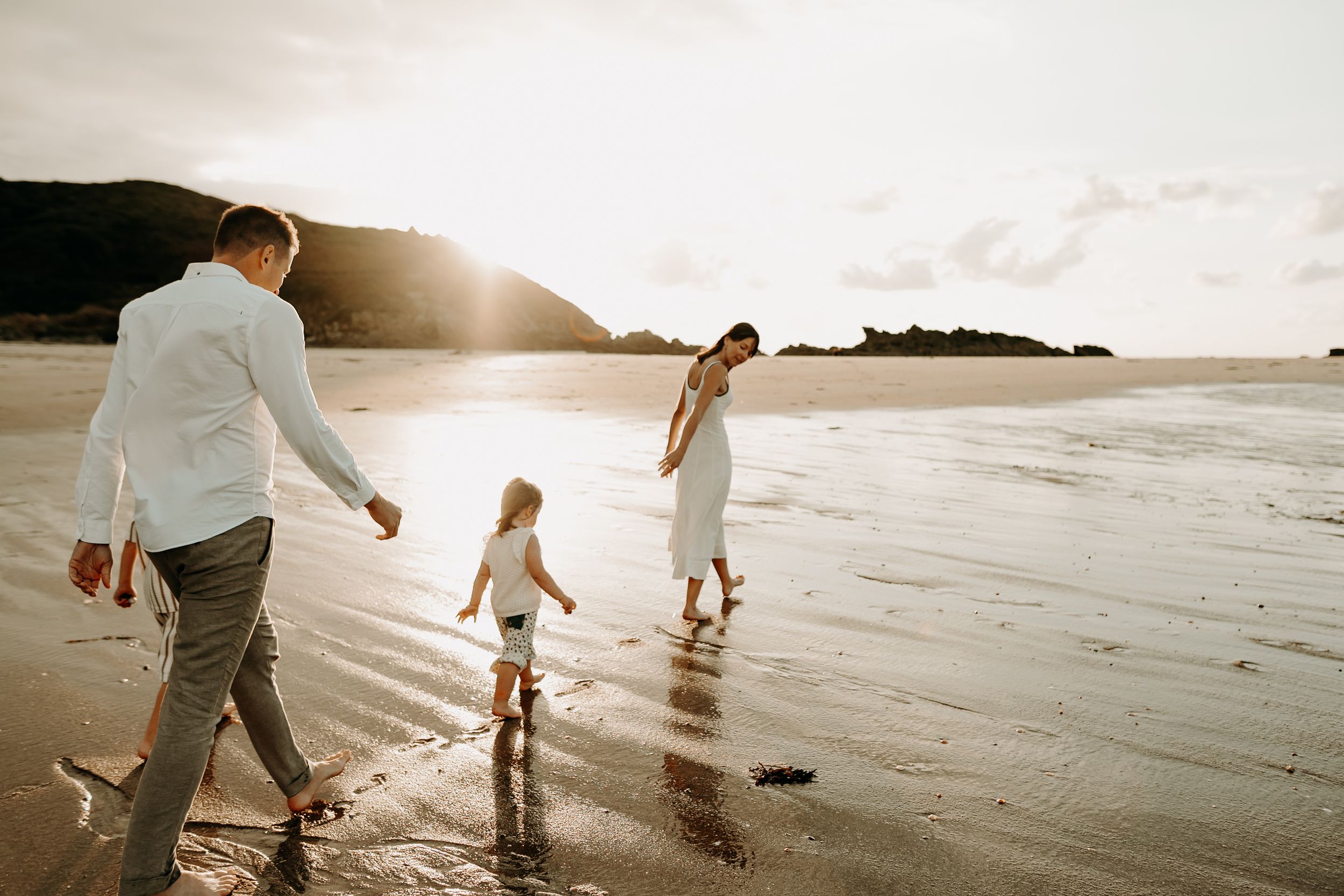 People walking along a scenic beach at sunset with sunlight streaming through clouds over the ocean.