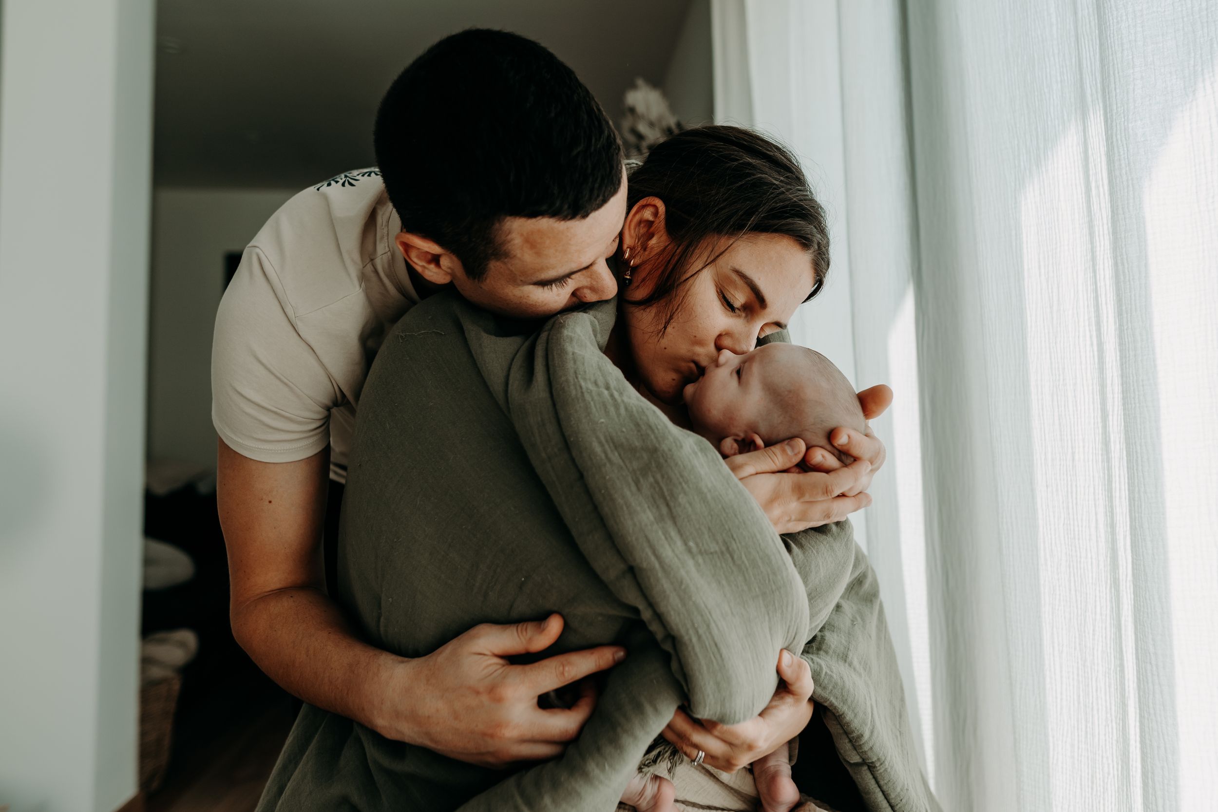 Tender moment between parents and baby while feeding from a bottle near a bright window.