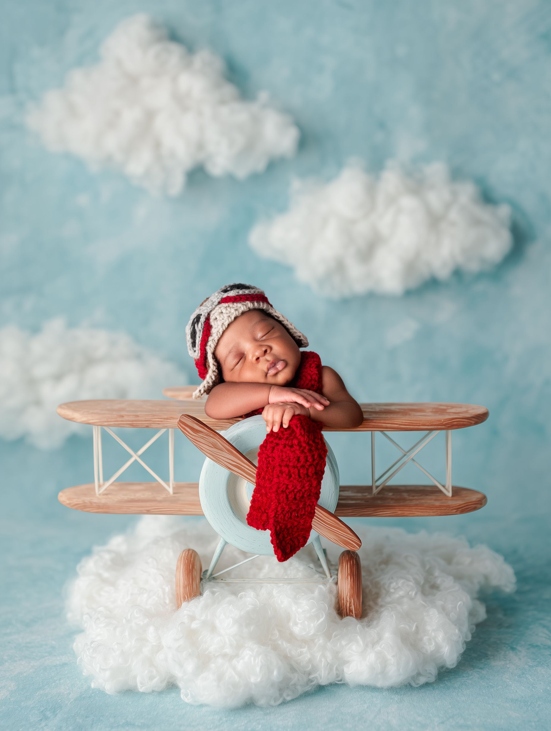 A sleeping baby in a red outfit lies on a wooden airplane prop against a cloudy blue backdrop.