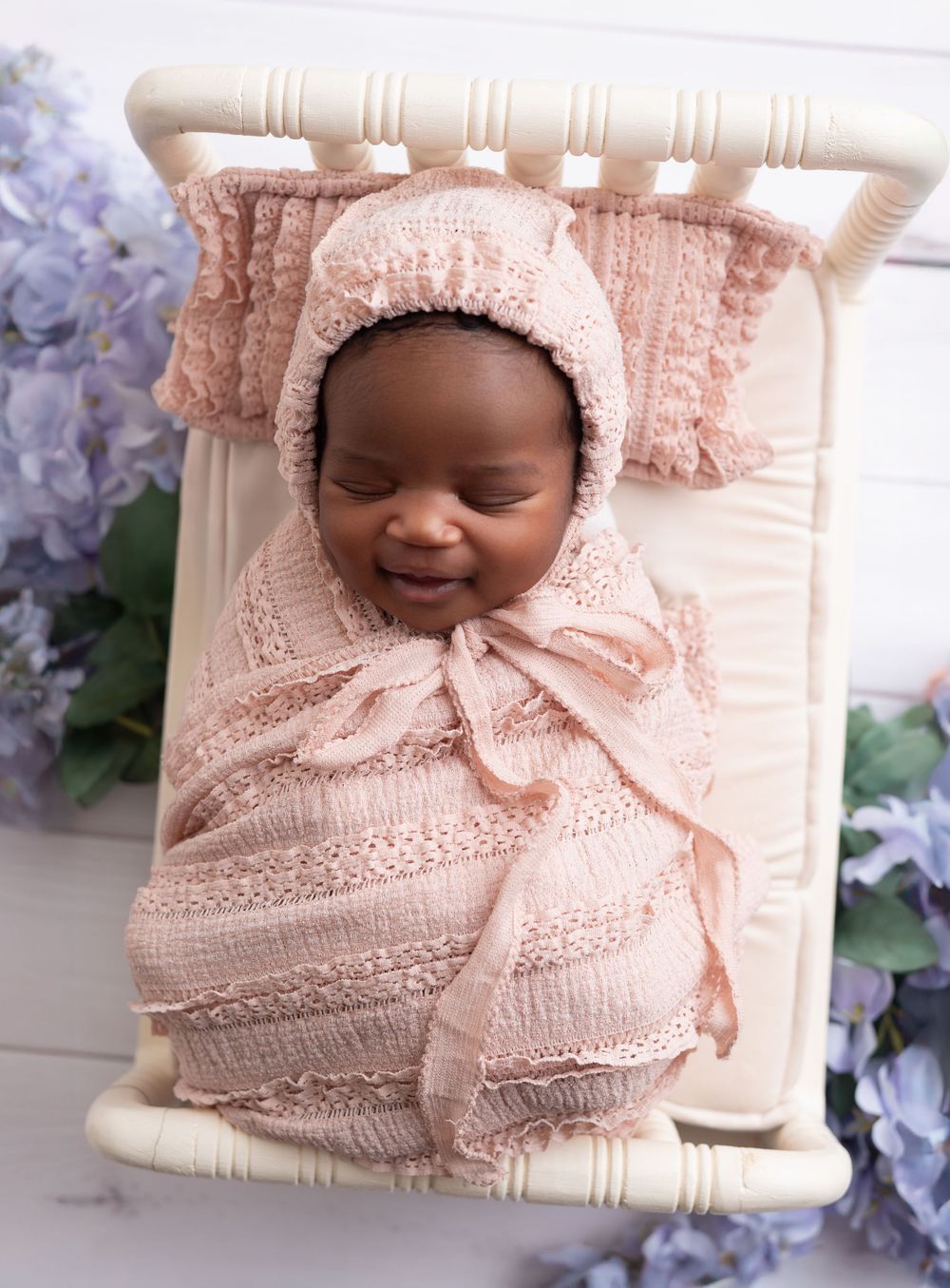 A delicate pink knit blanket with lace details is arranged with purple hydrangea flowers in a soft nursery setting.