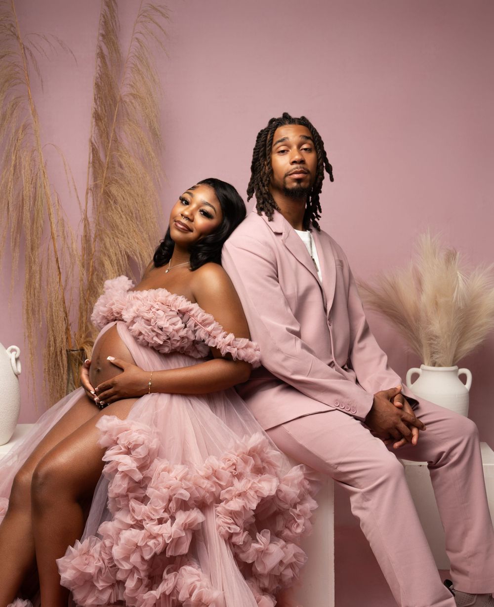 Couple in matching pink outfits posing for maternity shoot, woman in tulle gown, man in suit, pink studio backdrop.