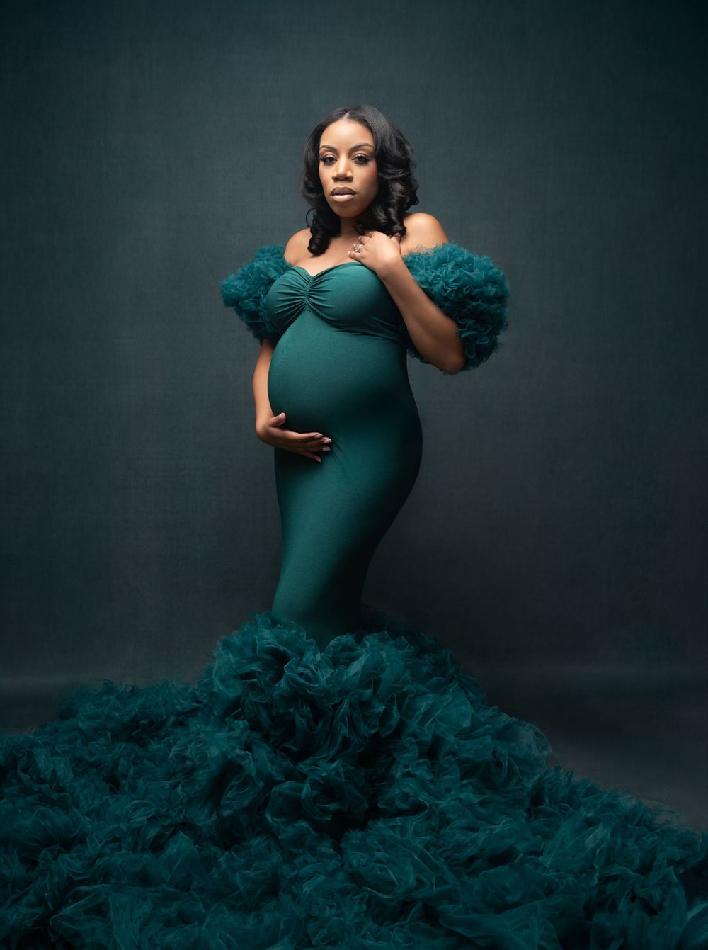 A figure in a dramatic emerald green gown with ruffled tulle details poses elegantly against a dark studio backdrop.