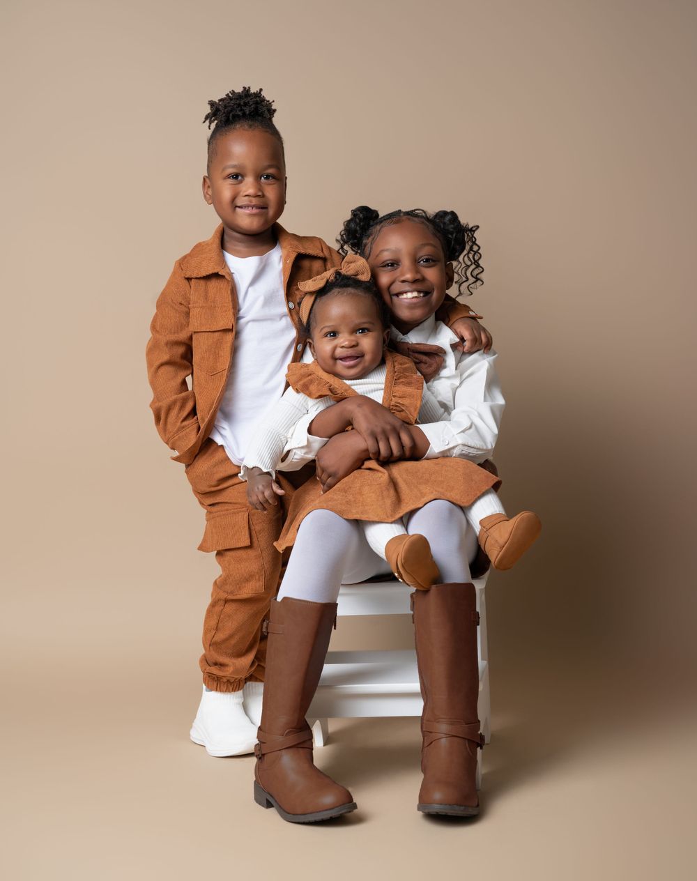 Toddler little kid and big kid posing for their sibling photoshoot in front of a neutral tan backdrop in matching white and brown outfits.